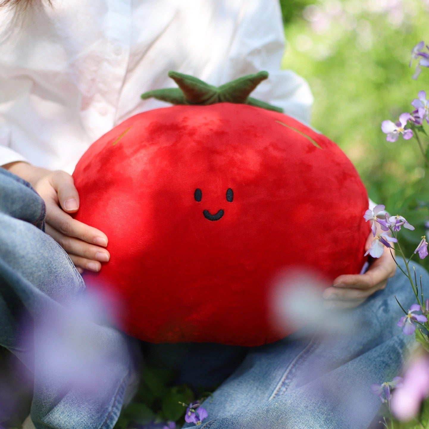 Person holding a plush tomato pillow in a natural setting with greenery and flowers.
