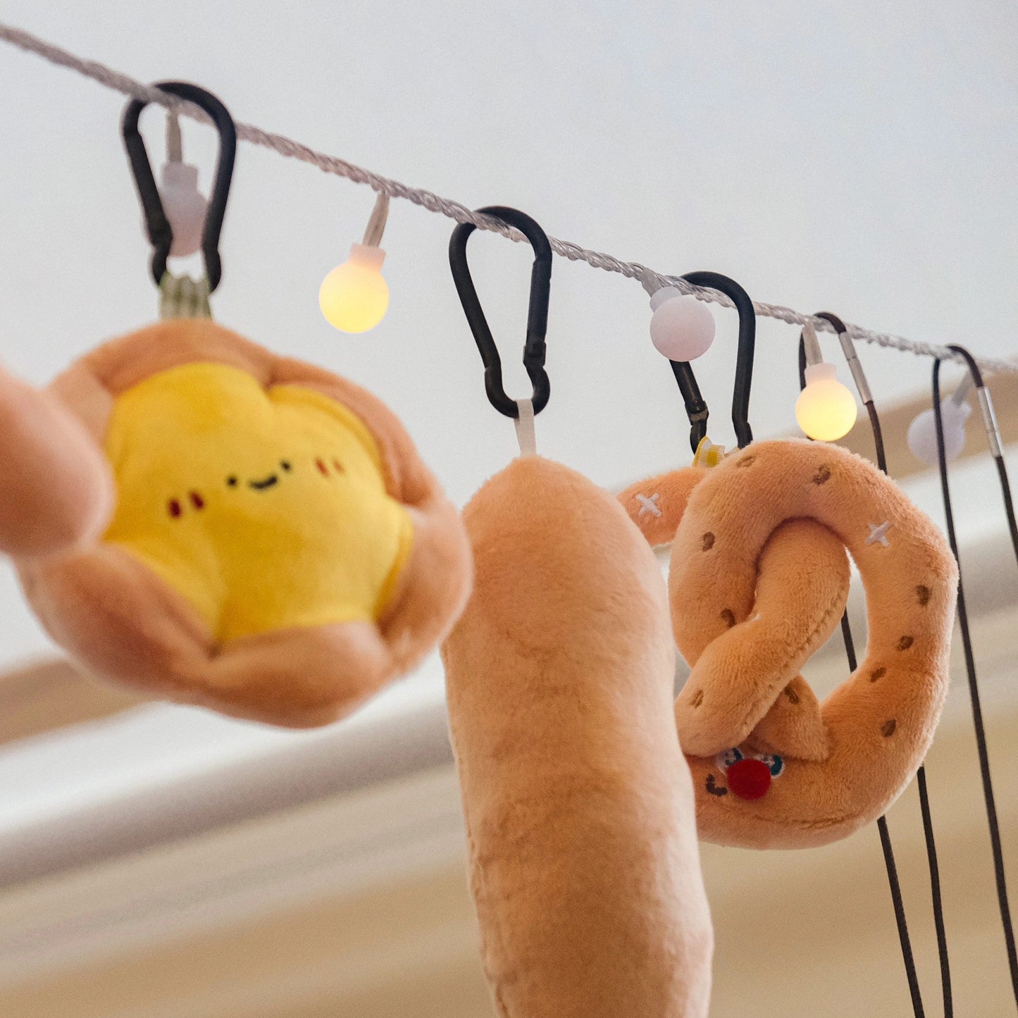Plush keychains shaped like a yellow star, brown bread roll, and orange pretzel on a string with lights.