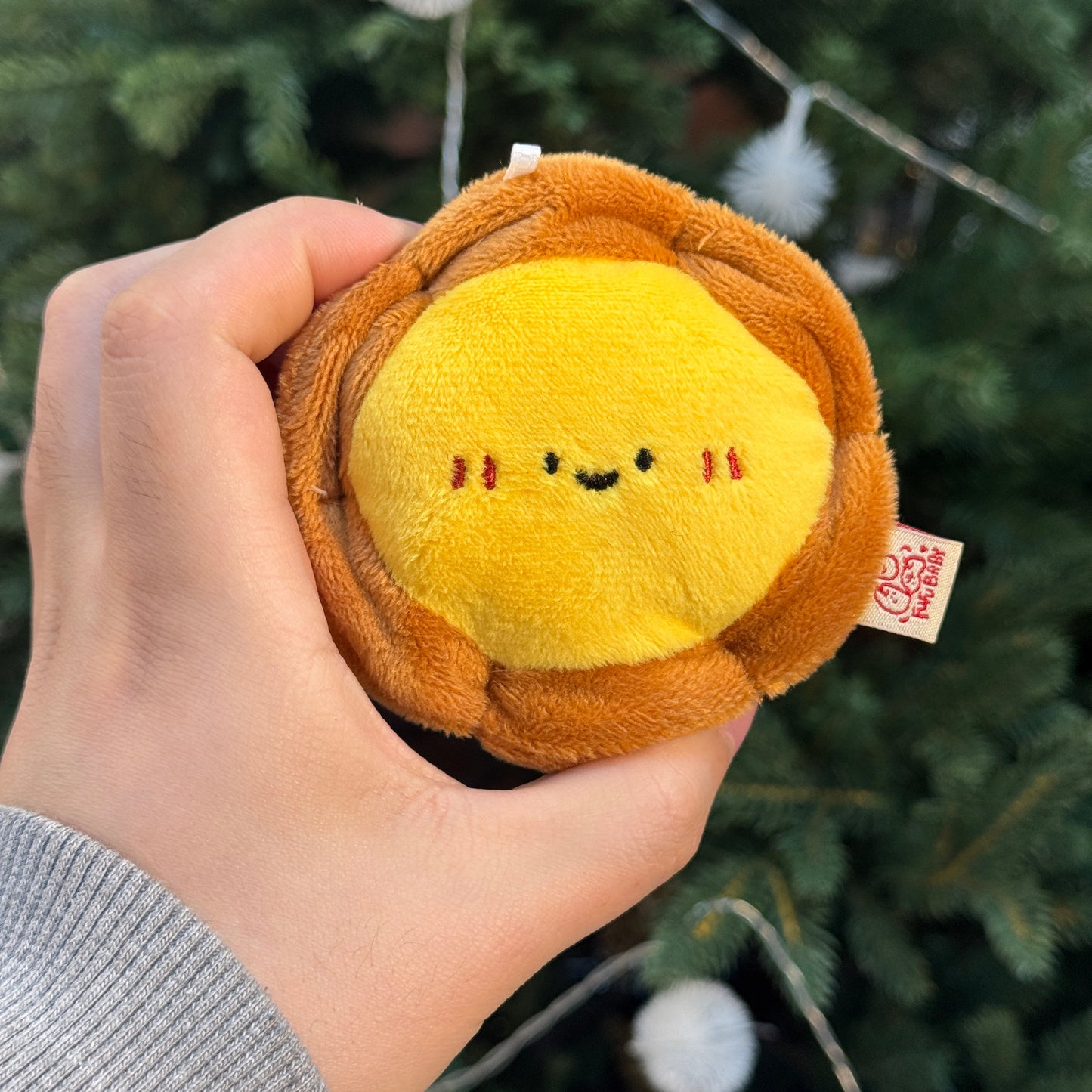 Hand holding a small plush lion toy with a Christmas tree in the background
