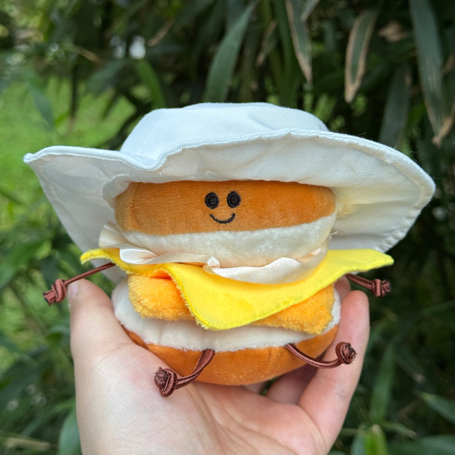 Hand holding a plush toy resembling a sandwich with a face on a green leafy background
